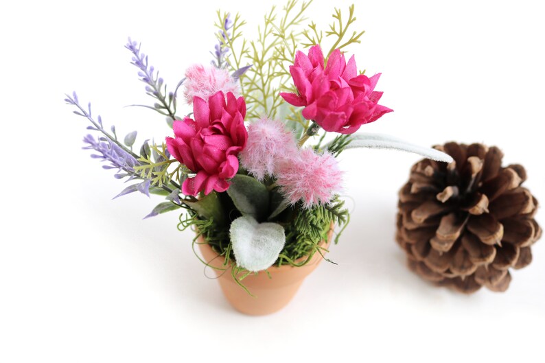 May include: A small terracotta pot filled with a floral arrangement featuring pink and purple flowers, green foliage, and white fuzzy accents. The pot is sitting on a white surface.