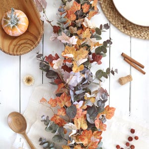 Pode incluir: Uma guirlanda decorativa de outono com uma mistura de folhas de bordo laranja, amarelas e castanhas intercaladas com folhagem de eucalipto verde ba&ccedil;o, disposta verticalmente sobre uma superf&iacute;cie branca junto a uma ab&oacute;bora e uma colher.