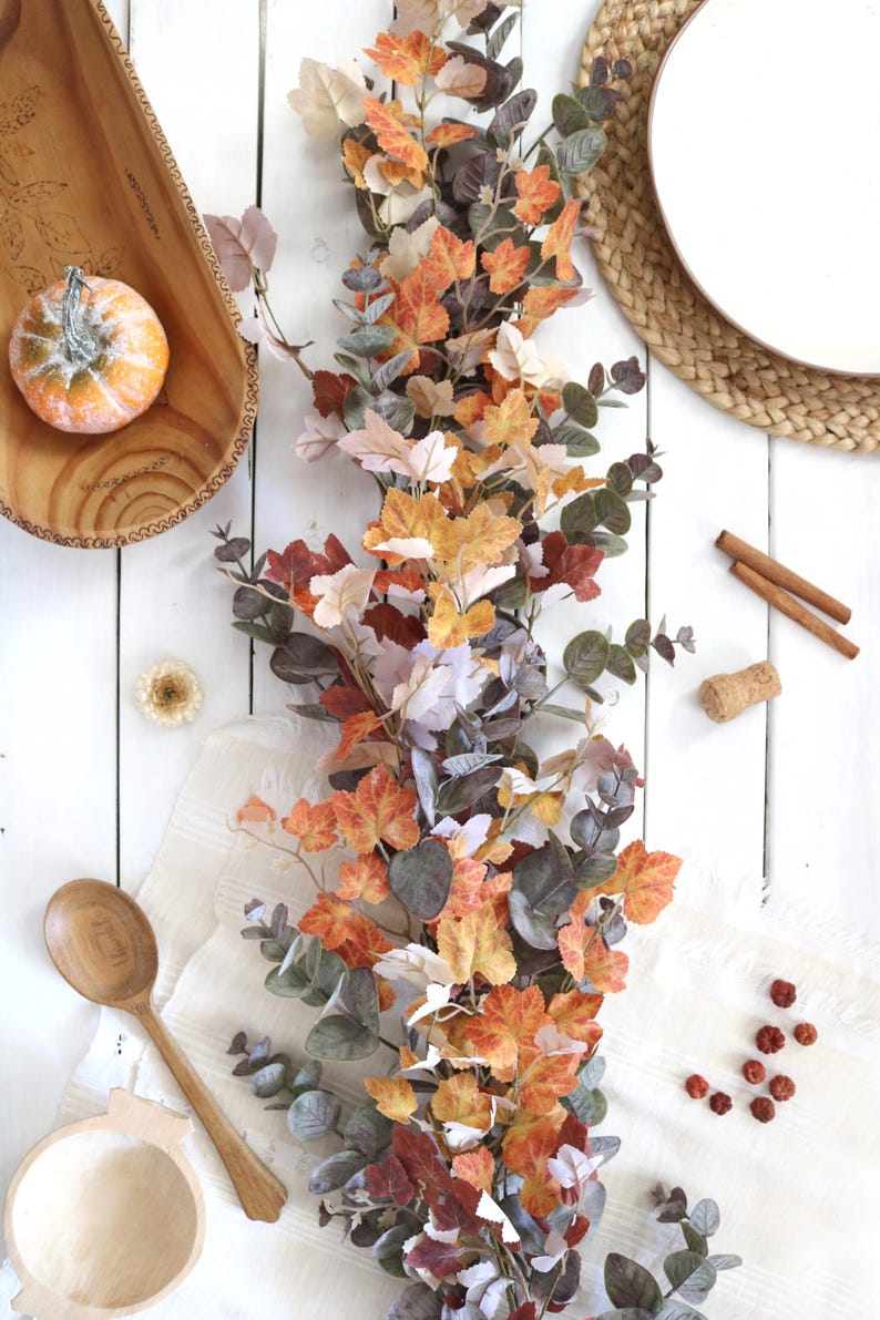 Pode incluir: Uma guirlanda decorativa de outono com uma mistura de folhas de bordo laranja, amarelas e castanhas intercaladas com folhagem de eucalipto verde ba&ccedil;o, disposta verticalmente sobre uma superf&iacute;cie branca junto a uma ab&oacute;bora e uma colher.