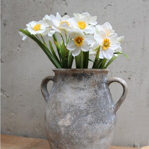 Op de afbeelding: Een rustieke, grijze vaas met twee handvatten bevat een boeket witte narcissen met gele centra en groene stelen. De vaas staat op een houten oppervlak tegen een getextureerde grijze muur.
