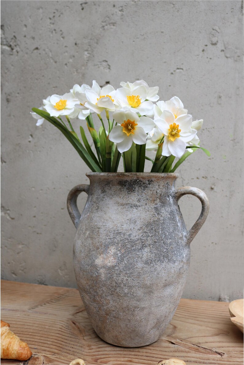Op de afbeelding: Een rustieke, grijze vaas met twee handvatten bevat een boeket witte narcissen met gele centra en groene stelen. De vaas staat op een houten oppervlak tegen een getextureerde grijze muur.