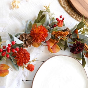 May include: A decorative garland featuring artificial orange and dark red dahlia flowers, green lamb's ear leaves, brown pinecones, and clusters of bright red and orange berries. The arrangement is composed of various faux botanical elements.