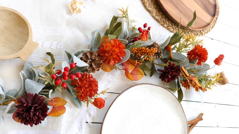 May include: A decorative garland featuring artificial orange and dark red dahlia flowers, green lamb's ear leaves, brown pinecones, and clusters of bright red and orange berries. The arrangement is composed of various faux botanical elements.