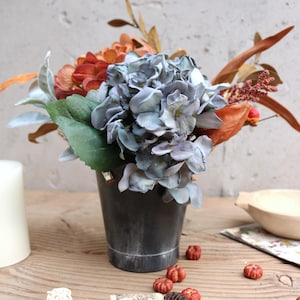 May include: A floral arrangement featuring blue and orange hydrangea blossoms, green leaves, and brown foliage housed in a dark gray metal bucket. The arrangement is surrounded by small orange pumpkins, pinecones, and a cork on a wooden surface.