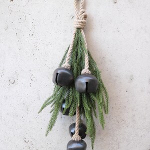 May include: A rustic Christmas decoration featuring four dark gray bells hanging from a natural rope and green pine branches. The bells are of varying sizes and are suspended from a knotted rope, creating a festive display.