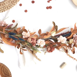 May include: A fall-themed floral garland with a variety of dried flowers and leaves in shades of brown, orange, and cream. The garland includes accents of blue and green, creating a rustic and natural aesthetic. Perfect for table decor.