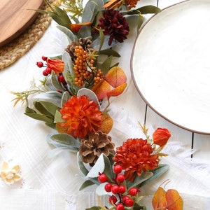 May include: A decorative autumn garland featuring orange and dark red chrysanthemum flowers, pinecones, clusters of bright red berries, orange berry sprigs, and soft, sage green lamb's ear leaves arranged in a long, flowing botanical display.