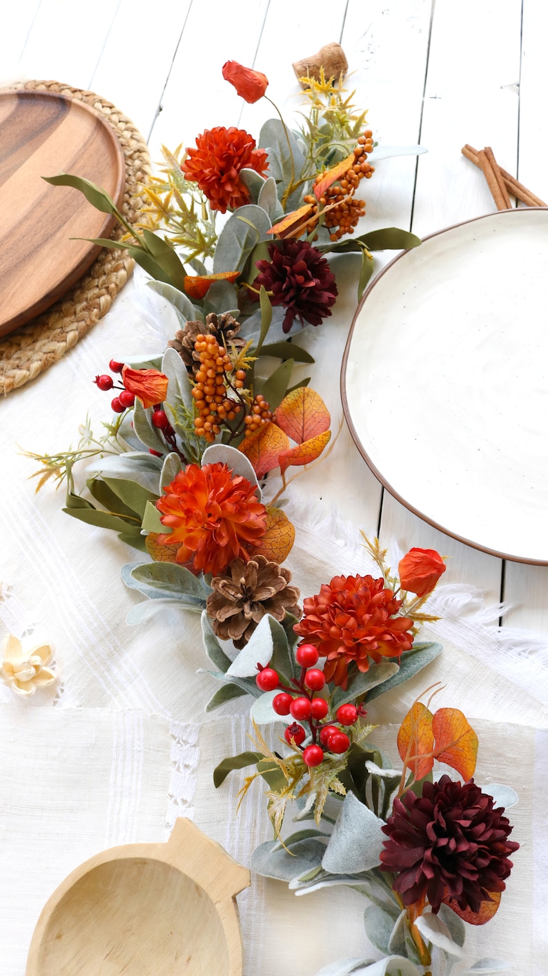 May include: A decorative autumn garland featuring orange and dark red chrysanthemum flowers, pinecones, clusters of bright red berries, orange berry sprigs, and soft, sage green lamb's ear leaves arranged in a long, flowing botanical display.