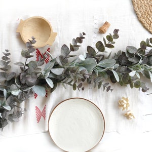 May include: A white ceramic plate with a light gray rim sits on a white surface with a garland of green and gray eucalyptus leaves. The garland is on a white surface with a red and white patterned fabric underneath.