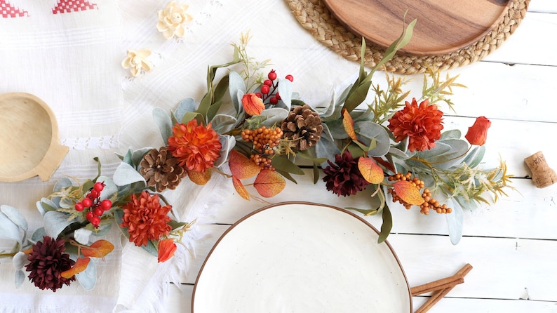 May include: A decorative autumn garland featuring artificial orange and dark red chrysanthemum flowers, brown pinecones, red berries, orange berry clusters, and sage green lamb's ear leaves arranged on a flexible vine base for seasonal display.