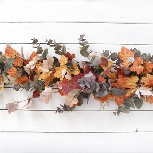 Pode incluir: Uma guirlanda decorativa com uma mistura de folhas de bordo artificiais em cores de outono, em tons de laranja, amarelo e vermelho, intercaladas com ramos de eucalipto verde suave. A folhagem est&aacute; disposta numa estrutura longa e rasteira.