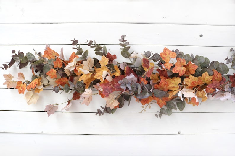 Pode incluir: Uma guirlanda decorativa com uma mistura de folhas de bordo artificiais em cores de outono, em tons de laranja, amarelo e vermelho, intercaladas com ramos de eucalipto verde suave. A folhagem est&aacute; disposta numa estrutura longa e rasteira.