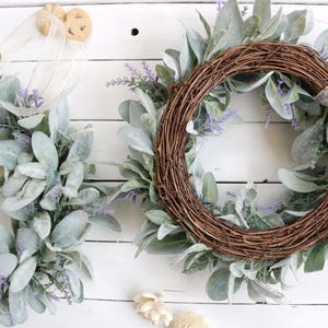 May include: Two wreaths with a rustic aesthetic. One is a full circle, the other is a half-circle. Both are made of woven brown twigs and adorned with large, soft, gray-green leaves and small purple flowers. The background is a white wooden surface.