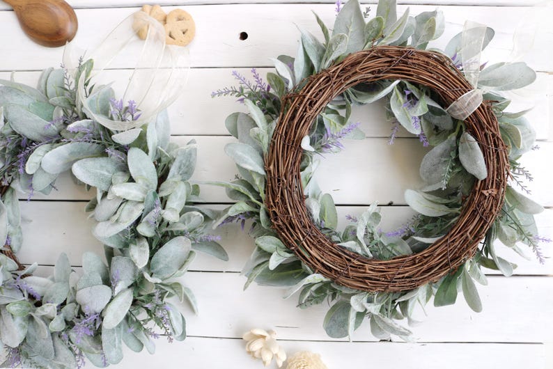 May include: Two wreaths with a rustic aesthetic. One is a full circle, the other is a half-circle. Both are made of woven brown twigs and adorned with large, soft, gray-green leaves and small purple flowers. The background is a white wooden surface.