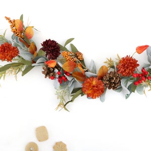 May include: A decorative garland featuring artificial orange and dark red chrysanthemums, pinecones, bright red berries, and sage green leaves. The arrangement includes small orange berry clusters and light green foliage, creating a seasonal display.