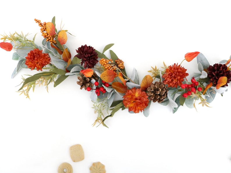 May include: A decorative garland featuring artificial orange and dark red chrysanthemums, pinecones, bright red berries, and sage green leaves. The arrangement includes small orange berry clusters and light green foliage, creating a seasonal display.