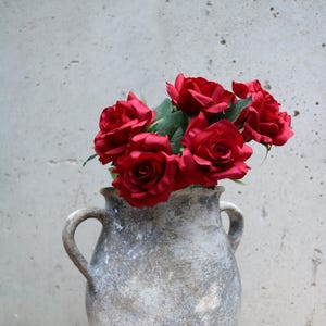 May include: A display of deep red roses with green foliage in a distressed grey ceramic vase. The vase features two loop handles and a textured finish, set against a plain backdrop.