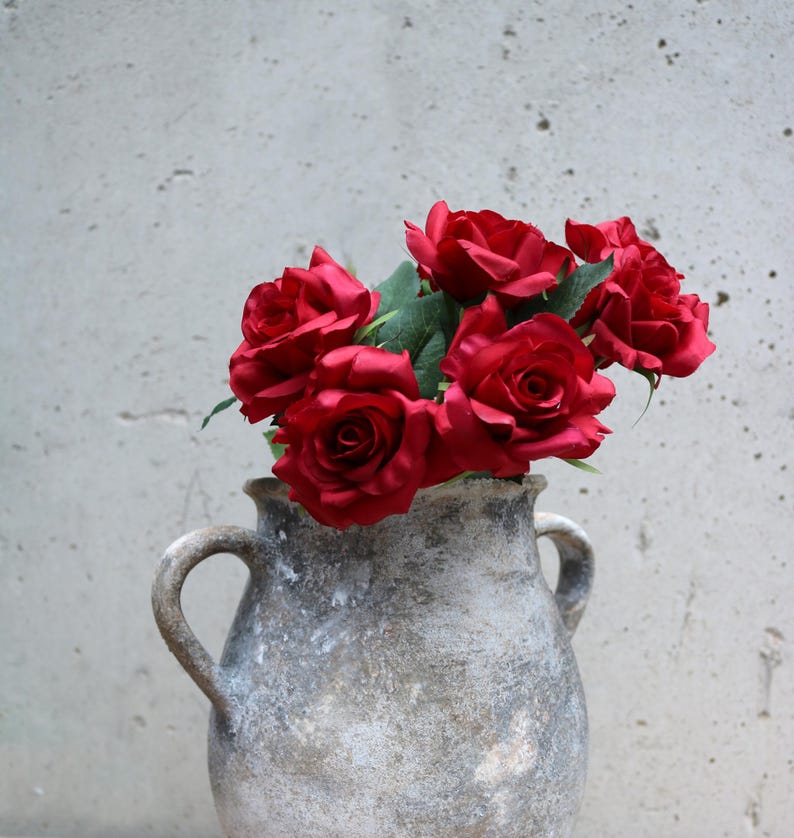 May include: A display of deep red roses with green foliage in a distressed grey ceramic vase. The vase features two loop handles and a textured finish, set against a plain backdrop.
