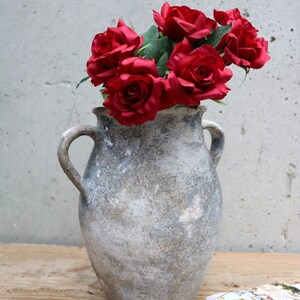 May include: A weathered, grey vase with two handles, holding a bouquet of deep red roses. The vase has a textured finish and is placed on a wooden surface. The roses provide a vivid contrast to the vase's muted tones.