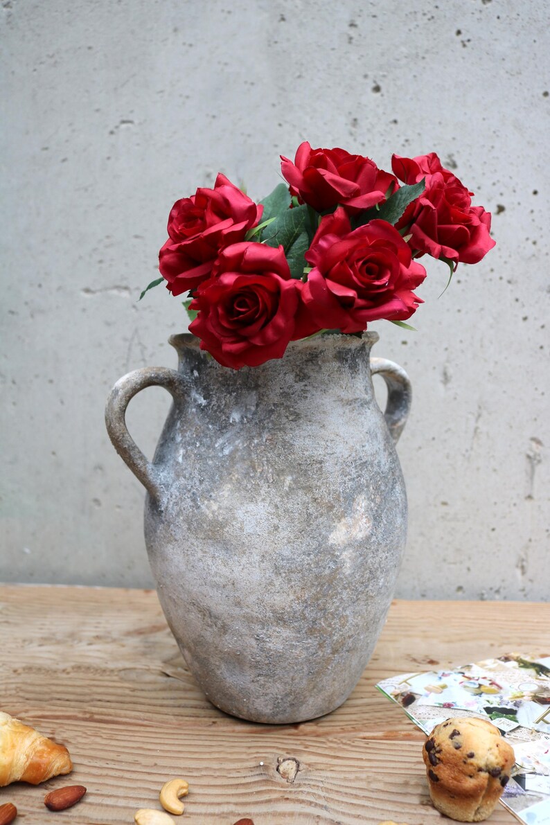 May include: A weathered, grey vase with two handles, holding a bouquet of deep red roses. The vase has a textured finish and is placed on a wooden surface. The roses provide a vivid contrast to the vase's muted tones.