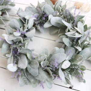 May include: A collection of decorative wreaths featuring large, soft, gray-green leaves and delicate purple flowers. The wreaths are arranged on a white wooden surface, creating a natural and elegant aesthetic. A small card with floral accents is also visible.