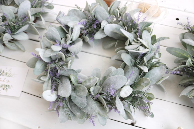 May include: A collection of decorative wreaths featuring large, soft, gray-green leaves and delicate purple flowers. The wreaths are arranged on a white wooden surface, creating a natural and elegant aesthetic. A small card with floral accents is also visible.