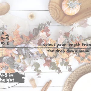 Pode incluir: Uma guirlanda com tema de outono apresentando uma mistura de folhas laranja, amarelas e marrons com ramos de eucalipto verde. A guirlanda mede 20-23 cm de largura e 10-13 cm de altura. O texto diz: select your length from the drop down menu.