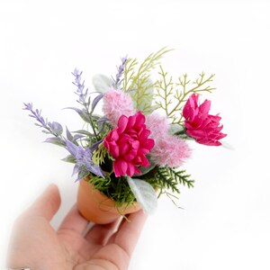 May include: A small terracotta pot filled with a floral arrangement. The arrangement includes pink flowers, lavender, and green foliage.
