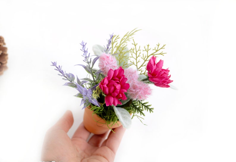May include: A small terracotta pot filled with a floral arrangement. The arrangement includes pink flowers, lavender, and green foliage.
