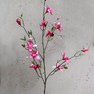 May include: A single stem of artificial magnolia flowers with vibrant pink blossoms and green leaves. The faux floral arrangement features several open flowers and closed buds, set against a neutral gray background.