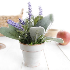 May include: A small white planter with a lavender and green plant arrangement. The planter has a distressed white finish and is sitting on a white surface.