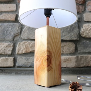 May include: A wooden table lamp with a white lampshade. The lamp base is a rectangular block of light-colored wood with visible grain. The lamp is on a gray surface in front of a stone wall.