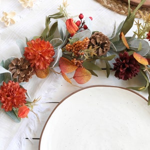 May include: A decorative garland featuring artificial orange and dark red chrysanthemums, pinecones, red berries, and sage green lamb's ear leaves. The arrangement includes small orange pods and brown berry clusters, creating a textured botanical display.