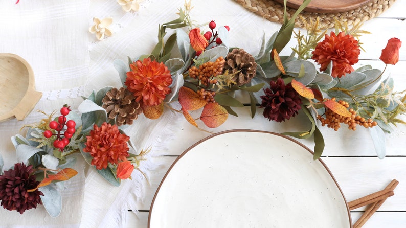 May include: A decorative garland featuring artificial orange and dark red chrysanthemums, pinecones, red berries, and sage green lamb's ear leaves. The arrangement includes small orange pods and brown berry clusters, creating a textured botanical display.