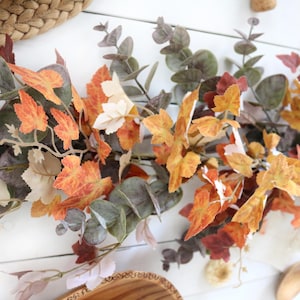 Pode incluir: Uma guirlanda decorativa com uma mistura de folhas de eucalipto artificiais em tons de verde e cinza suaves, intercaladas com folhas de bordo vibrantes em cores de outono em tons de laranja, amarelo e vermelho profundo, dispostas em videira.