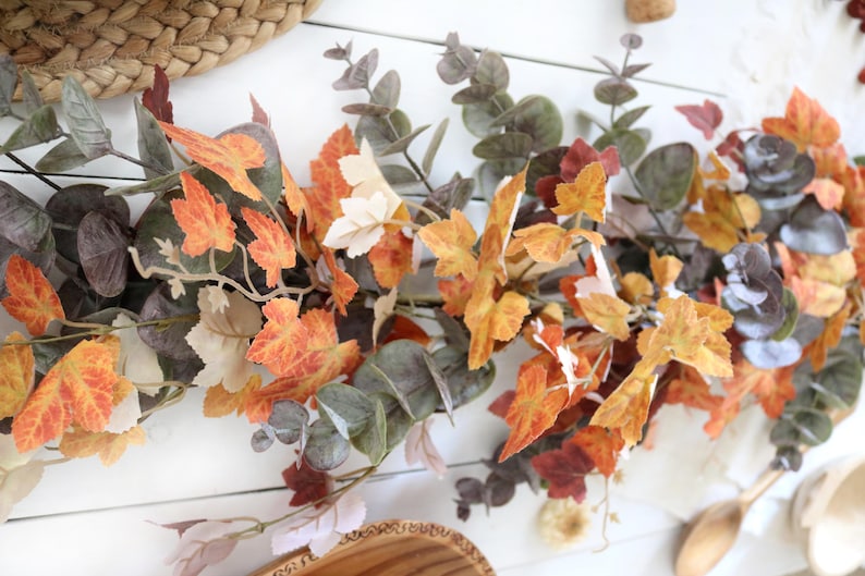 Pode incluir: Uma guirlanda decorativa com uma mistura de folhas de eucalipto artificiais em tons de verde e cinza suaves, intercaladas com folhas de bordo vibrantes em cores de outono em tons de laranja, amarelo e vermelho profundo, dispostas em videira.