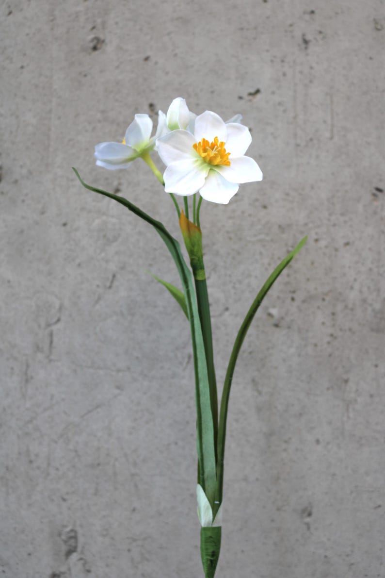 Op de afbeelding: Een enkele stengel van kunstmatige narcissen met witte bloemblaadjes en een heldergeel centrum. De bloem heeft groene bladeren en een groene stengel. De achtergrond is een effen, lichtgrijze muur.