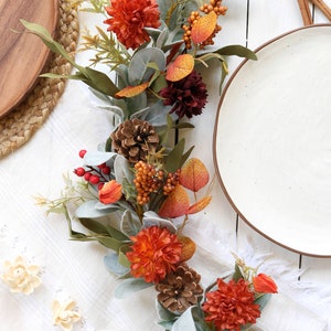 May include: An artificial autumn garland featuring orange and dark red chrysanthemum flowers, brown pinecones, red berries, orange berry clusters, and sage green lamb's ear leaves. The decorative floral arrangement is elongated and flexible.
