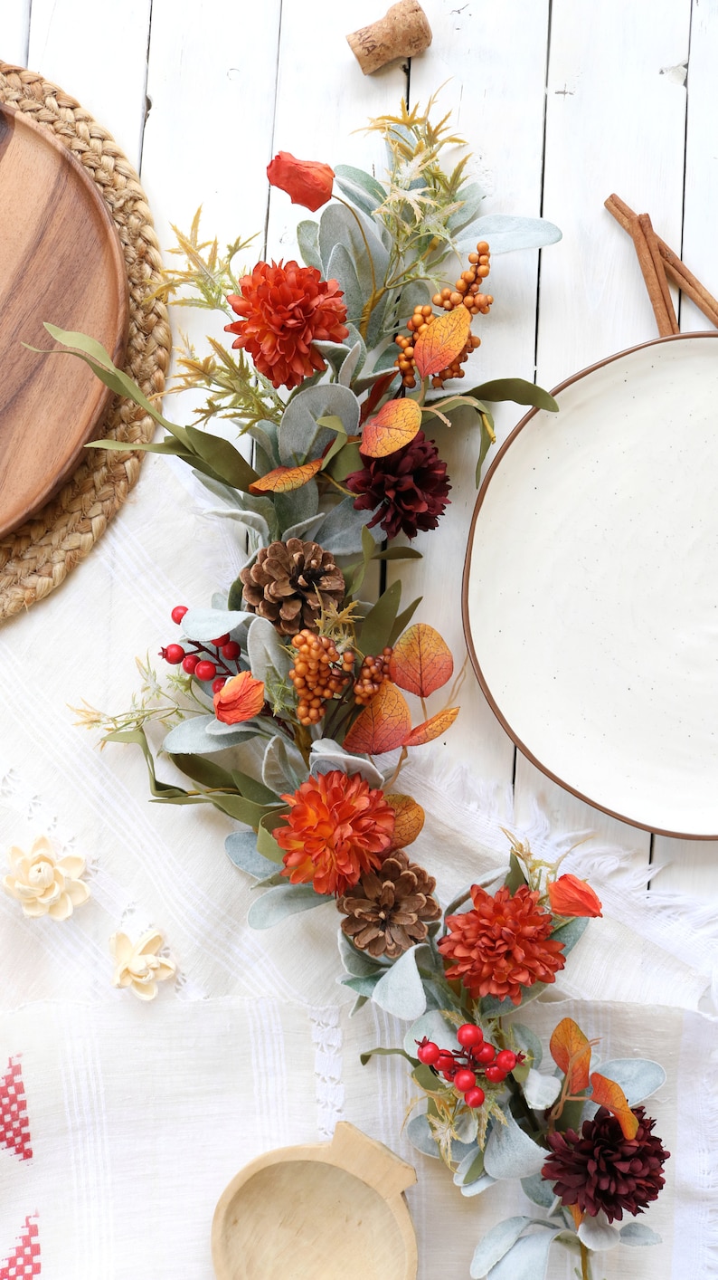 May include: An artificial autumn garland featuring orange and dark red chrysanthemum flowers, brown pinecones, red berries, orange berry clusters, and sage green lamb's ear leaves. The decorative floral arrangement is elongated and flexible.