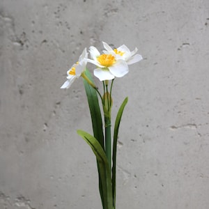 Op de afbeelding: Een enkele stengel van kunstmatige narcissen met witte bloemblaadjes en gele centra. De groene stengel heeft lange, slanke bladeren. De bloemen staan tegen een getextureerde grijze achtergrond.