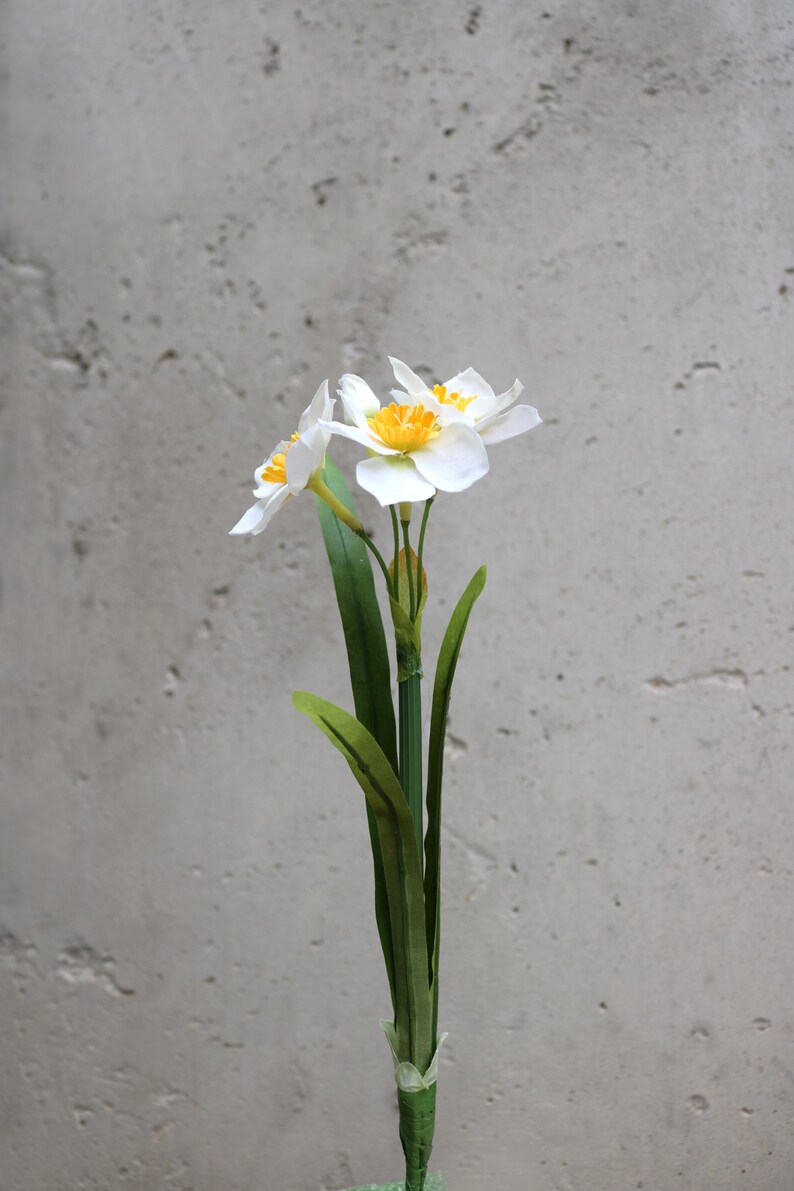 Op de afbeelding: Een enkele stengel van kunstmatige narcissen met witte bloemblaadjes en gele centra. De groene stengel heeft lange, slanke bladeren. De bloemen staan tegen een getextureerde grijze achtergrond.