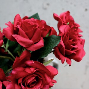 May include: A bunch of deep red roses with green foliage. The flowers are fully open, displaying their velvety petals. The arrangement is set against a plain backdrop, emphasising the rich colour of the roses.