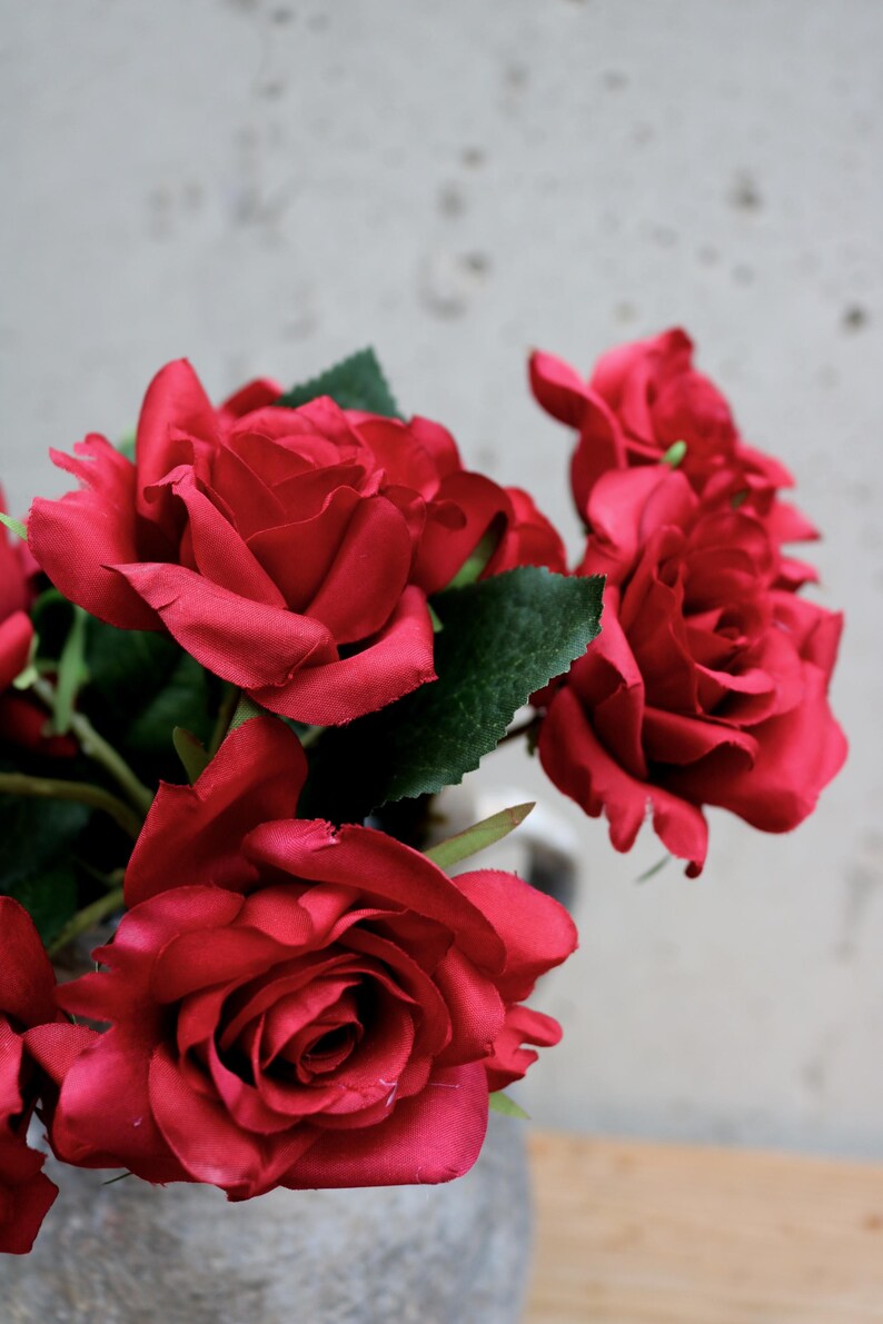 May include: A bunch of deep red roses with green foliage. The flowers are fully open, displaying their velvety petals. The arrangement is set against a plain backdrop, emphasising the rich colour of the roses.