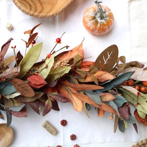 May include: An autumn-themed garland featuring a mix of faux foliage in shades of orange, brown, muted green, and deep red. The arrangement includes various leaf shapes, small orange berries, and wheat stalks, creating a dense, textured display.