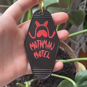 May include: Black motel key chain with red text that reads "Mothman Motel". The key chain features a red silhouette of a mothman with red eyes.