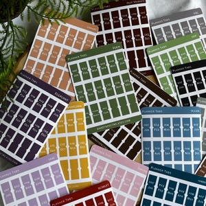 May include: A collection of rectangular sticker sheets featuring planner tabs in various solid colors including caramel, deep maroon, fern, eggplant, dandelion, lilac, sakura, ocean, and harbor. Each sheet contains months from January to December.