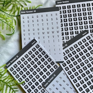 May include: A set of black and white alphabet and number stickers. The stickers are in a square shape with rounded corners. The stickers are arranged in rows and columns on a white background. The text on the stickers includes the letters A through Z and the numbers 1 through 9.