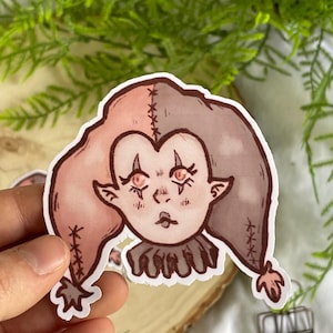 May include: A pink and brown cartoon jester with stitched seams and large eyes. The jester is wearing a ruffled collar.