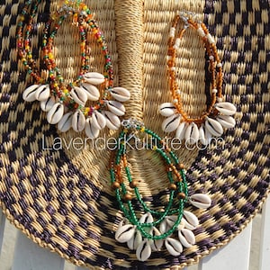 May include: Three beaded necklaces with cowrie shells. The necklaces are made with brown, orange, green, and white beads. The necklaces are displayed on a woven mat.