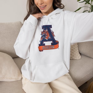 May include: A white hooded sweatshirt featuring a large navy blue letter A with an orange and black tiger graphic leaping through it. Below the letter is an orange banner with the word AUBURN in white text. The hoodie has long sleeves and a front pocket.